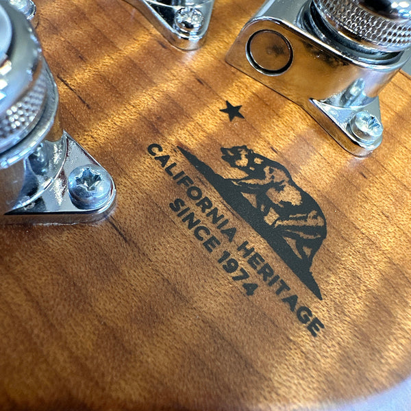 Close-up of California Heritage guitar logo on wood grain body with chrome hardware and tuning pegs visible