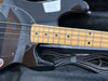 Close-up of black electric bass guitar with maple neck and fretboard, featuring four strings and black dot inlays, resting in a padded black case with a coiled black instrument cable.
