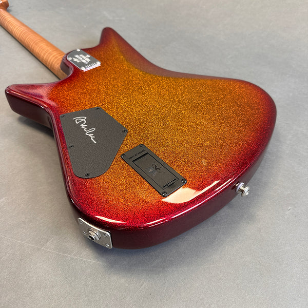 Electric guitar body with orange to red sparkle finish, showing rear control cavity covers and input jack, on a gray surface