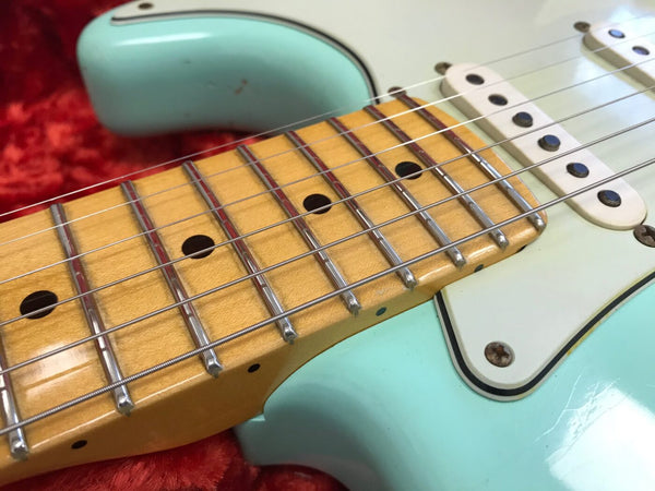 Close-up of light blue electric guitar with maple fretboard and white single-coil pickups on red textured background