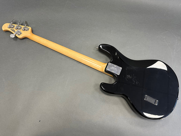 Black electric bass guitar with natural wood neck and chrome tuning pegs, shown from the back on gray background