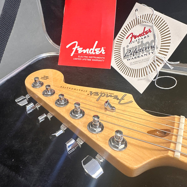 Close-up of Fender Stratocaster guitar headstock with tuning pegs and Fender lifetime warranty tags on black case interior