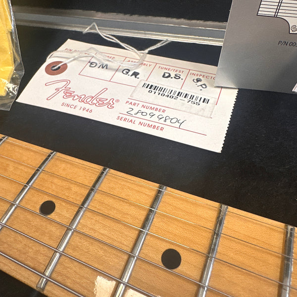 Close-up of maple guitar fretboard with metal frets and black dot inlays next to a Fender tag showing part number 28099804 and handwritten tuning and inspection initials.