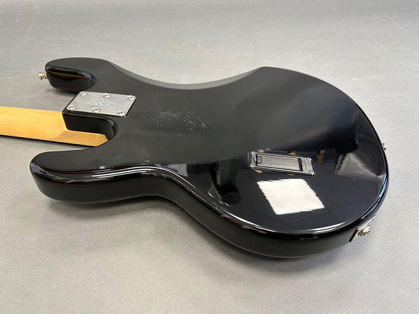 Back side view of a black electric guitar with a maple neck, showing chrome neck plate and battery compartment on a gray surface