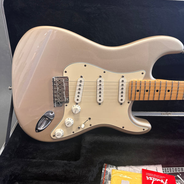 Close-up of metallic silver electric guitar with maple fretboard, three single-coil pickups, white pickguard, and tone controls in black plush-lined guitar case with Fender warranty card and yellow polishing cloth.
