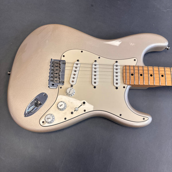 Close-up of metallic silver electric guitar body with white pickguard, three single-coil pickups, volume and tone control knobs, and maple neck on dark gray surface
