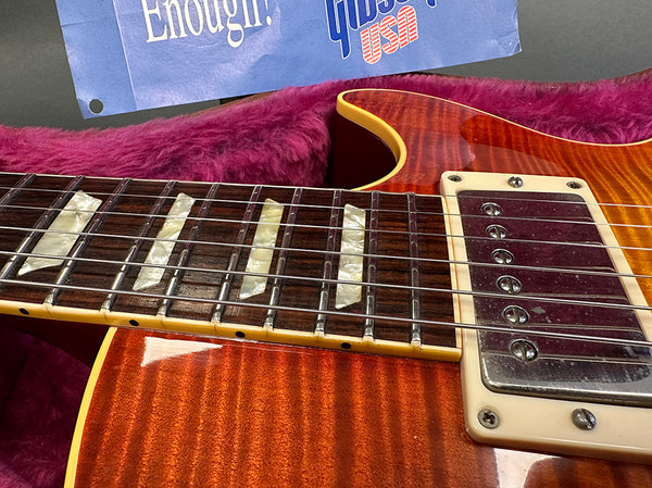 Close-up of sunburst electric guitar neck and pickups with pearl block inlays resting on pink plush guitar case lining, partially visible vintage Gibson USA poster in background