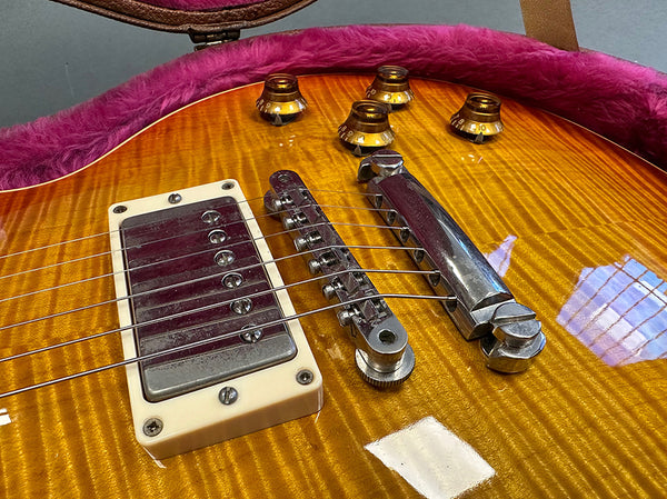 Close-up of honey burst electric guitar body showing humbucker pickup, Tune-o-matic bridge, chrome tailpiece, and amber control knobs in a guitar case with pink plush lining