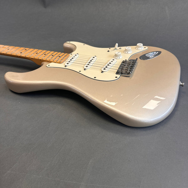 Close-up of metallic champagne electric guitar body with three single-coil pickups, white pickguard, tone and volume knobs, and maple neck on gray background