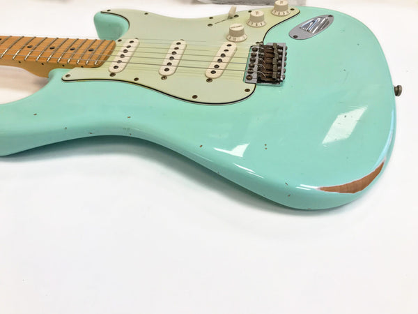 Close-up view of seafoam green electric guitar body with vintage white pickguard, three single-coil pickups, maple fretboard, and visible wear and paint chipping near the edges on white background