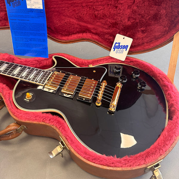 Black Gibson electric guitar with gold hardware and three pickups in a pink plush-lined hard case with Gibson USA tag and blue certificate