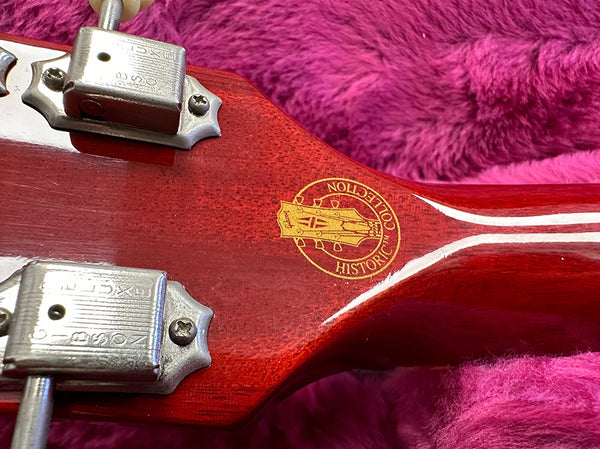 Close-up of the back of a guitar headstock with vintage tuning pegs and a "Historic Collection" logo on a polished red wood finish, resting on a plush pink surface.
