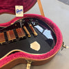 Close-up of black Gibson electric guitar with gold hardware and wooden pickup covers inside a plush pink-lined brown guitar case with Gibson USA tag