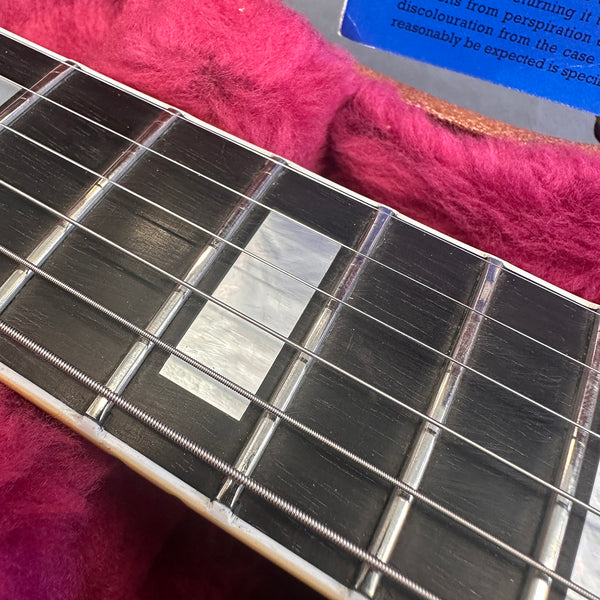Close-up of guitar fretboard showing mother of pearl rectangular inlays and steel strings on dark wood fingerboard, resting inside pink plush-lined guitar case.