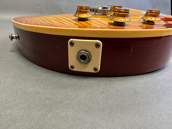Close-up of electric guitar jack input and amber control knobs on a flamed maple top with cream binding and dark cherry body finish.
