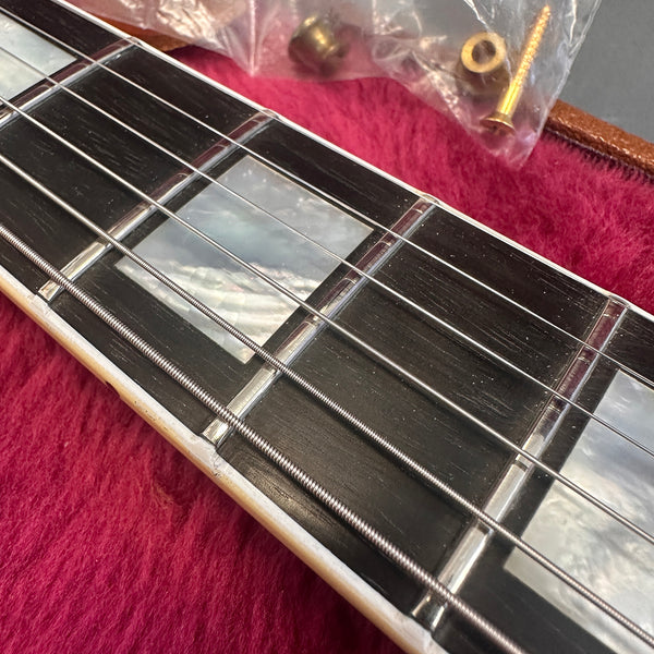 Close-up of guitar fretboard with mother of pearl block inlays, metal strings, and part of guitar neck resting on pink plush surface with brass screws in plastic bag background