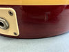 Close-up of maroon electric guitar body showing input jack and cream-colored plastic plate with minor surface dent and scratches on grey background.