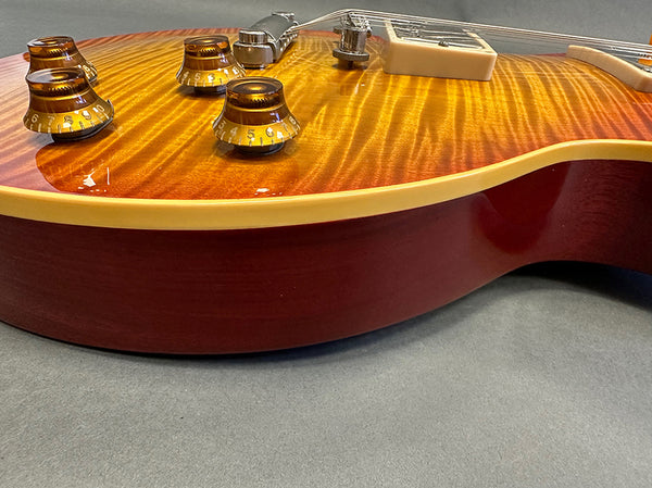 Close-up of electric guitar body with sunburst finish, amber control knobs, cream binding, and chrome bridge on gray surface