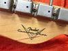 Close-up of Fender Custom Shop logo stamped on light wood guitar headstock with vintage tuning pegs and red fabric background