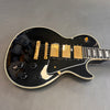 Black electric guitar with gold pickups and hardware, black control knobs, and white binding on body and fretboard on gray background