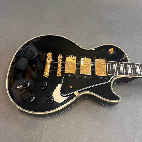 Black electric guitar with gold pickups and hardware, black control knobs, and white binding on body and fretboard on gray background
