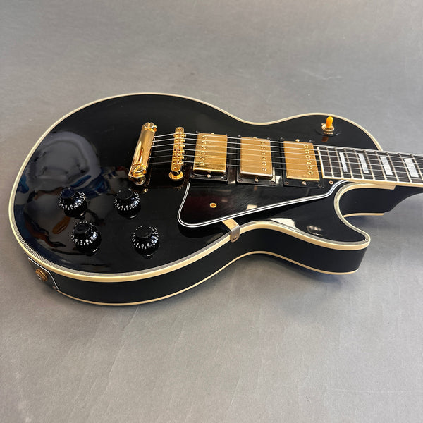 Black electric guitar with gold hardware, three humbucker pickups, four control knobs, and cream binding on body edges, displayed on gray surface