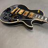 Black electric guitar with gold hardware and control knobs on gray surface, single cutaway body style.