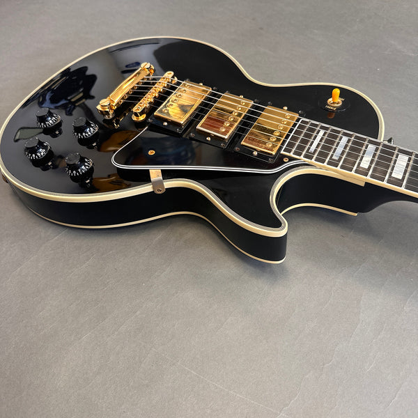 Black electric guitar with gold hardware and control knobs on gray surface, single cutaway body style.