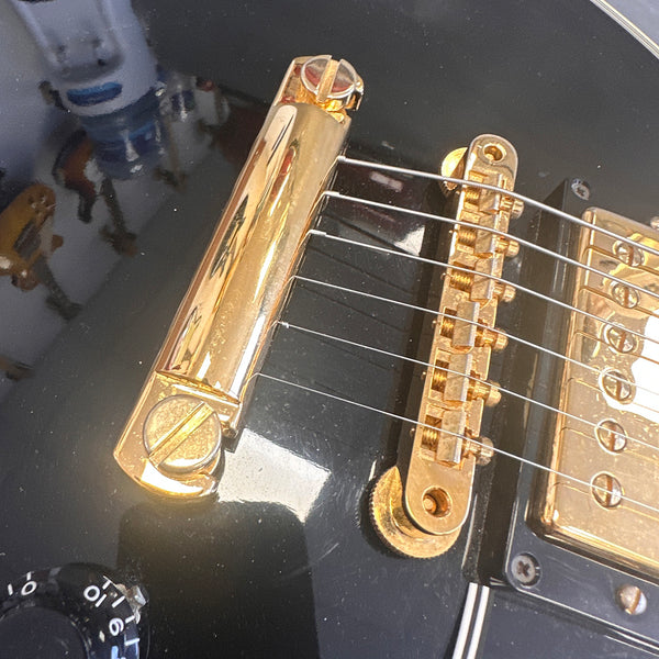 Close-up of black electric guitar bridge and tailpiece with gold hardware and six strings, showing volume knob and pickup details