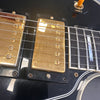 Close-up of black electric guitar body showing gold humbucker pickups, strings, and pickguard detail with treble switch.