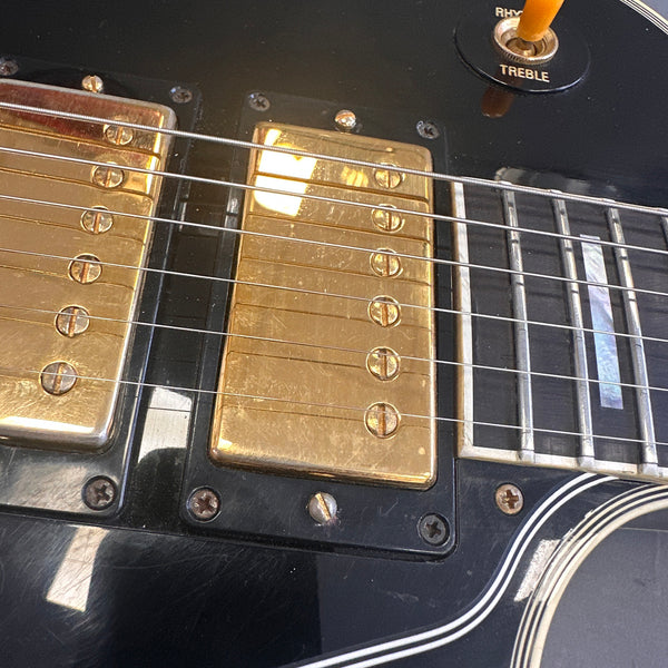 Close-up of black electric guitar body showing gold humbucker pickups, strings, and pickguard detail with treble switch.