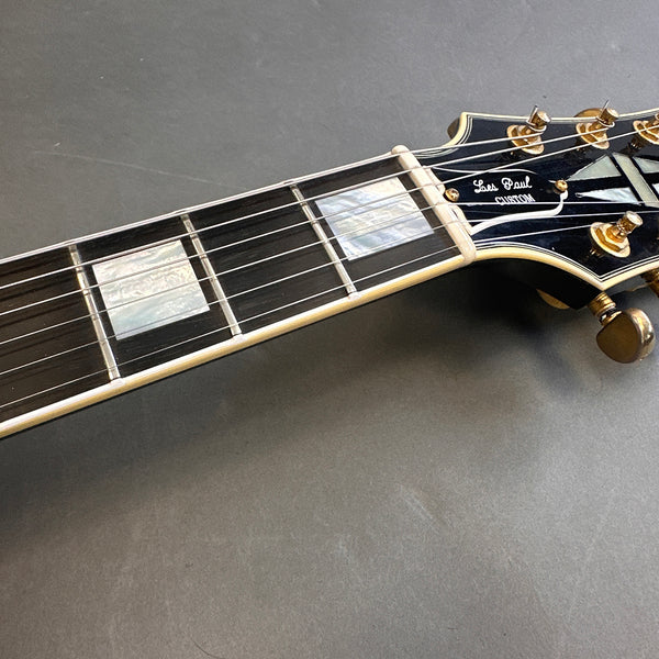 Close-up of Les Paul Custom guitar headstock and fretboard with mother of pearl inlays and gold tuning pegs on dark background