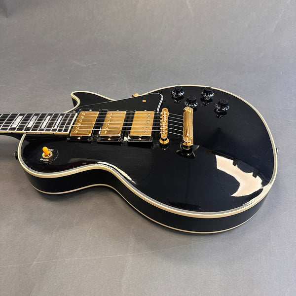 Black electric guitar with gold pickups and hardware, white binding, and black control knobs on grey background.