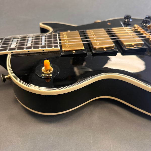 Close-up of black electric guitar body with cream binding, three gold humbucker pickups, and an orange rhythm/treble toggle switch on grey background.
