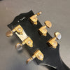 Close-up of black guitar headstock with gold Grover tuning pegs and serial number 7 7061 on gray background.