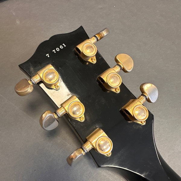 Close-up of black guitar headstock with gold Grover tuning pegs and serial number 7 7061 on gray background.