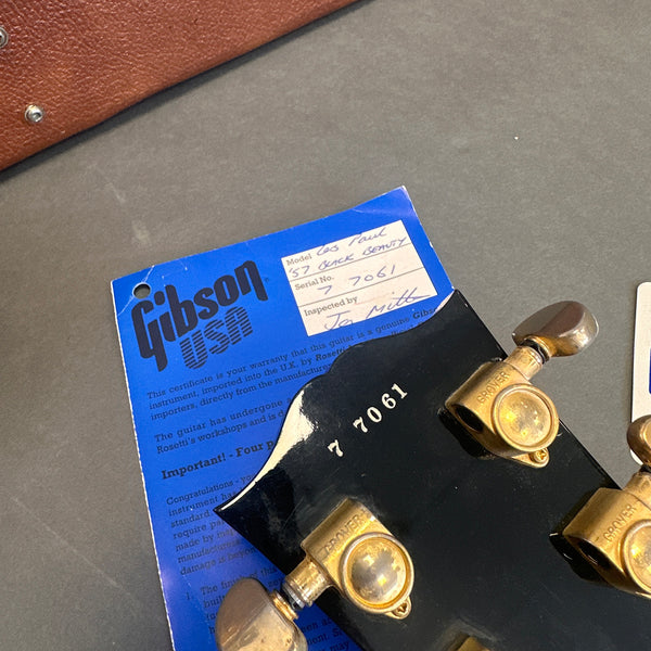 Close-up of Gibson USA blue warranty card and black guitar headstock with serial number 77061 and gold Grover tuning pegs for Gibson Les Paul '57 Buck Beauty model