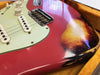 Close-up of vintage electric guitar body with red sunburst finish, white pickguard, single-coil pickups, and volume and tone control knobs, showing wear and aging in yellow-lined guitar case
