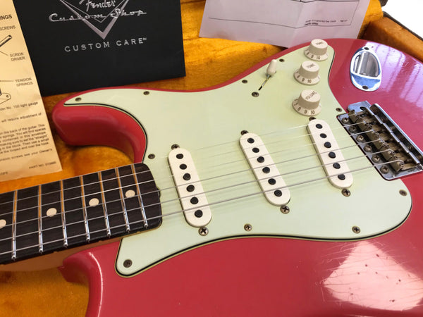 Close-up of vintage red Fender Stratocaster electric guitar with white pickguard, three single-coil pickups, and tone and volume knobs inside a yellow-lined guitar case with Fender Custom Shop paperwork and setup instructions visible.