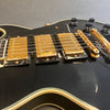 Close-up of black electric guitar body with gold pickup covers, gold bridge, and black control knobs with white numbering