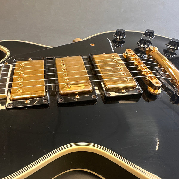 Close-up of black electric guitar body with gold pickup covers, gold bridge, and black control knobs with white numbering