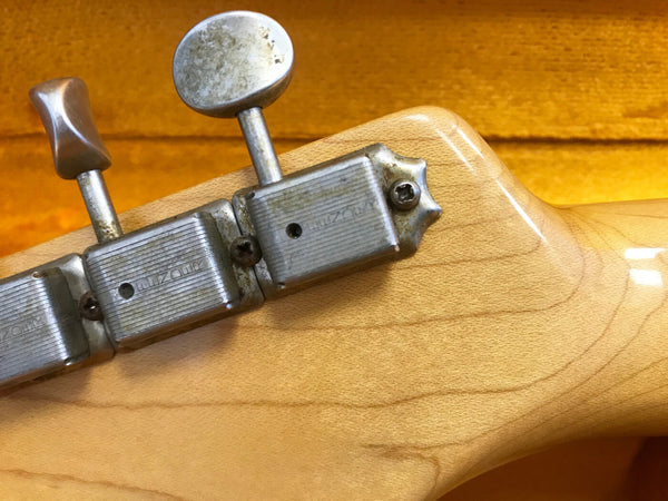 Close-up of vintage MIZON tuning pegs on the back of a light wood guitar headstock against a yellow background
