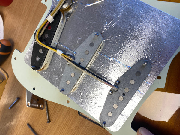 Close-up of three single-coil electric guitar pickups mounted on a pickguard with shielding foil, showing wiring and screws for installation on a wooden workbench.