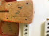 Close-up of guitar neck heel with handwritten signature and markings, showing wood grain texture and screw holes, next to white guitar body with pickup covers and screws.