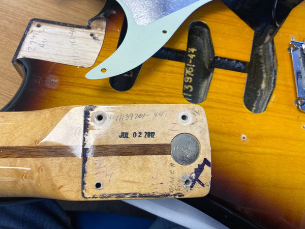 Close-up of a vintage sunburst electric guitar body and detached neck showing neck pocket with handwritten dates and stamp markings, including "JUL 02 2012" and serial numbers.