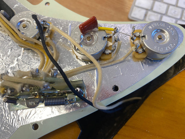 Close-up of electric guitar control plate showing three potentiometers, capacitor, wiring, and spring tension mechanism on metallic shielded pickguard.