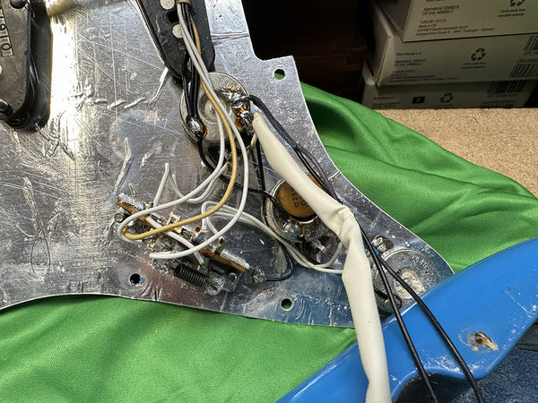 Close-up of guitar pickguard electronics showing wiring, potentiometers, and capacitor components on silver metal plate with green cloth background and blue guitar body edge visible.