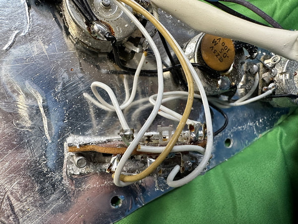 Close-up of vintage electric guitar wiring and potentiometers showing white and yellow wires soldered to metal control plate with green fabric background