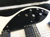Close-up of white electric guitar with black pickguard, control knobs, toggle switch, and bridge in guitar case with plush black interior.