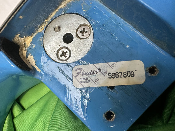 Close-up of blue Fender guitar body showing metal plate with two screws and a Fender serial number sticker S967809 on worn painted wood surface